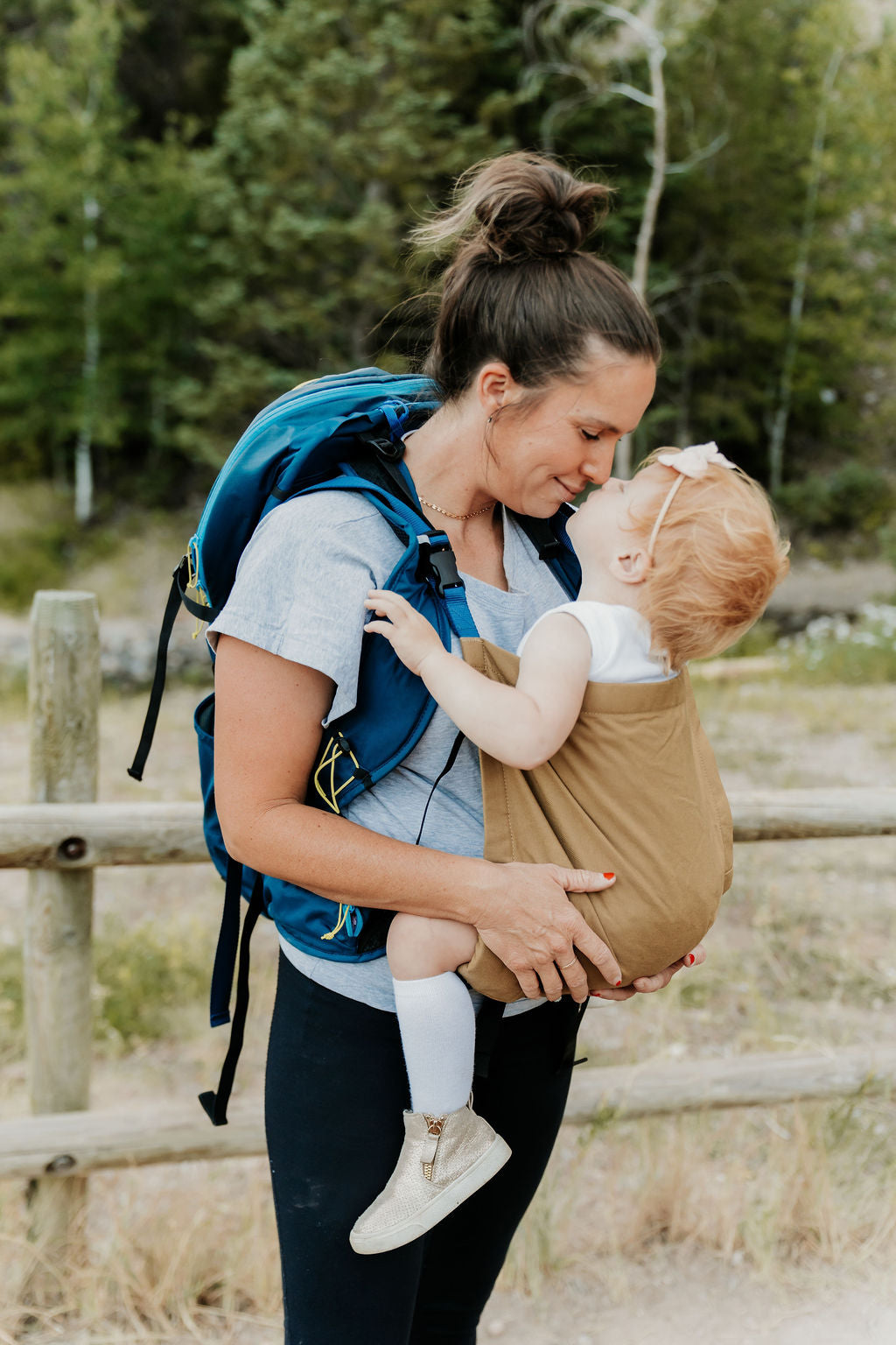 Woman carrying a child in a sling and wearing a blue backpack in a forest setting