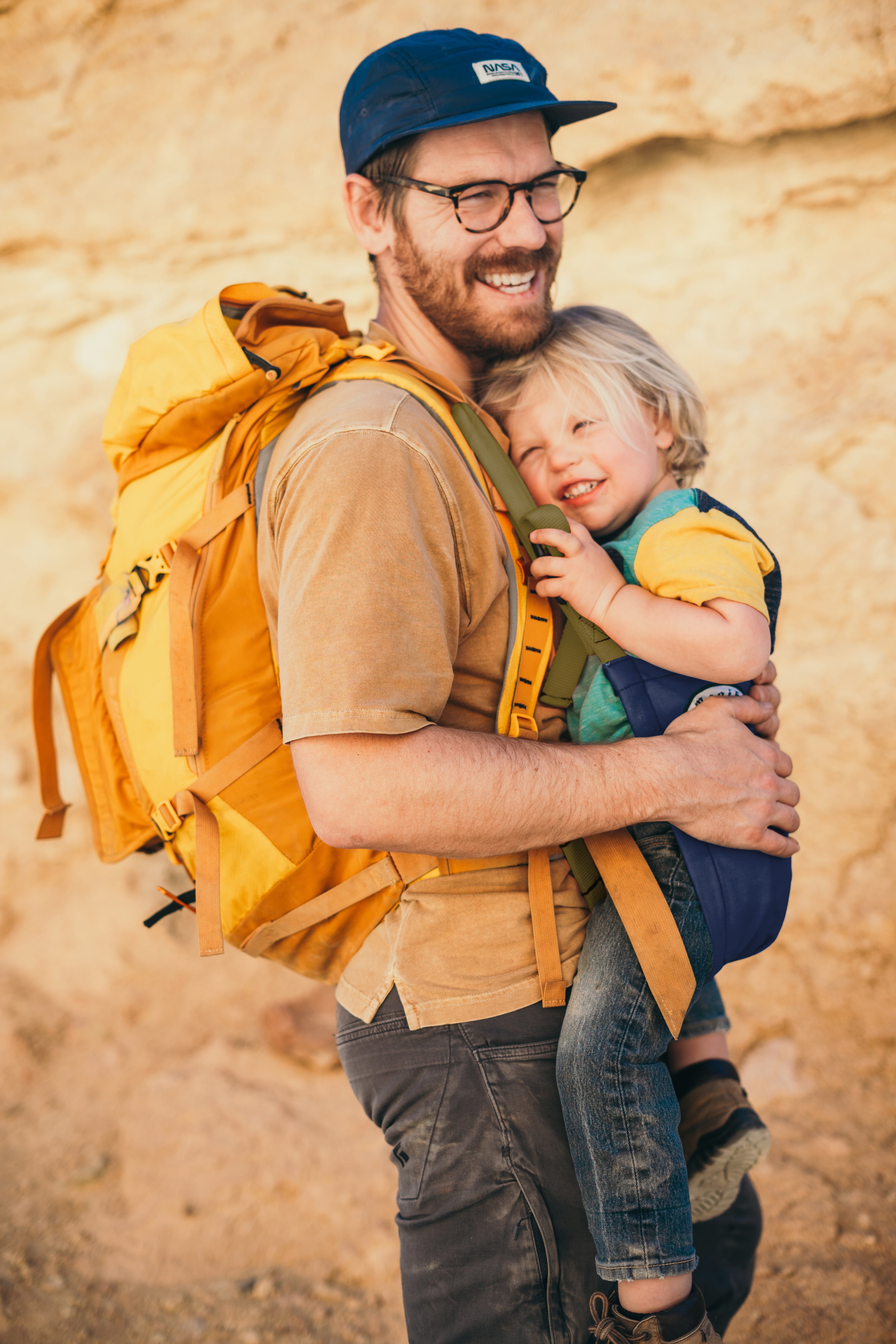 Man with a yellow backpack holding a child against a rocky background