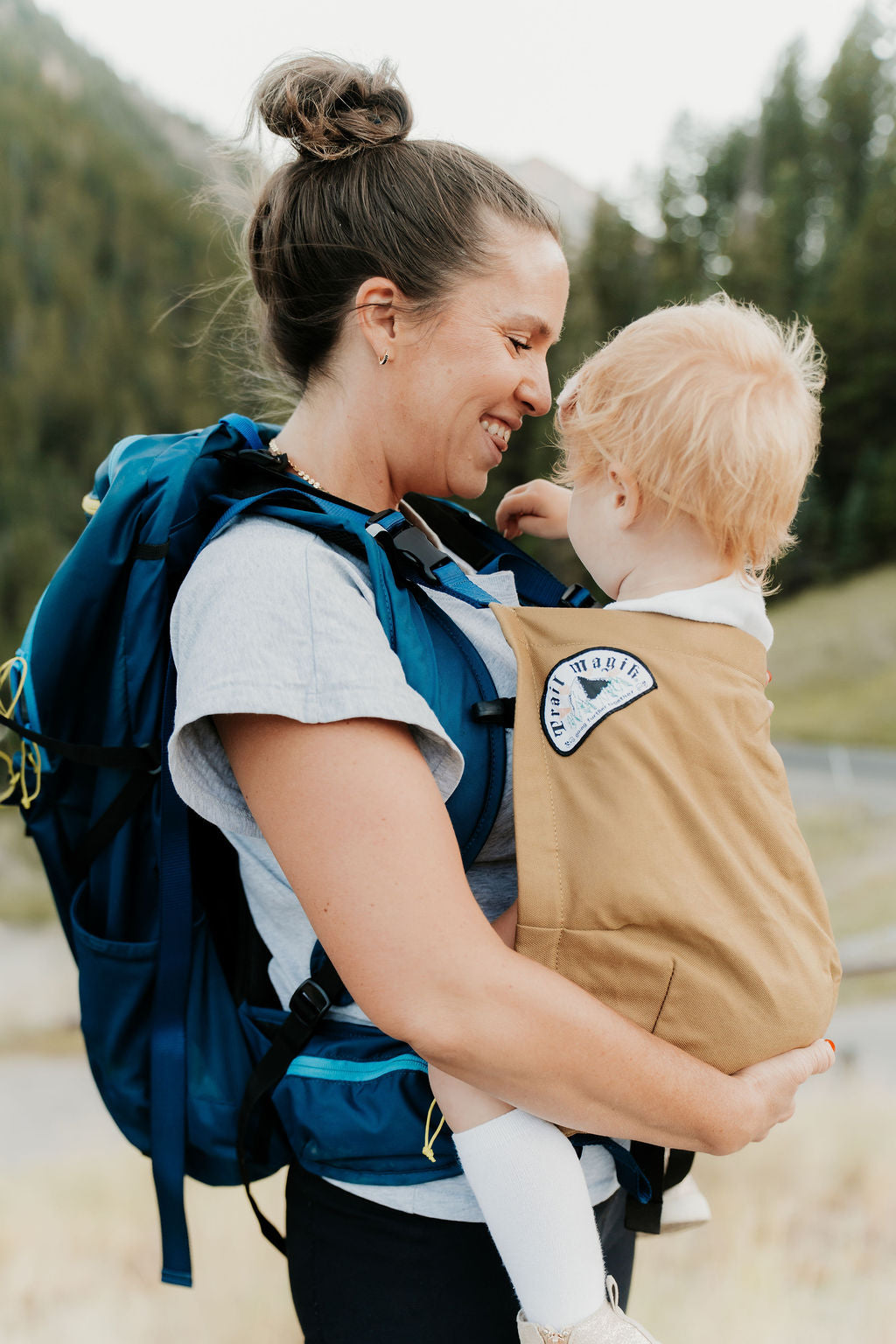 Woman carrying a child in a baby carrier with a blue backpack outdoors.