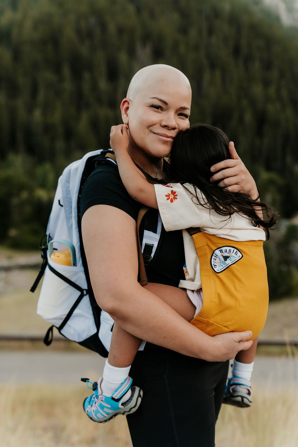 Woman carrying a child in a baby carrier outdoors with greenery in the background