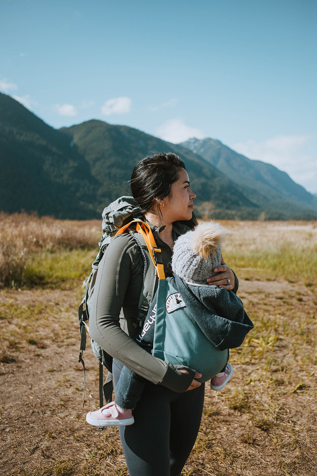 Woman with a baby in a carrier, standing in a field with mountains in the background