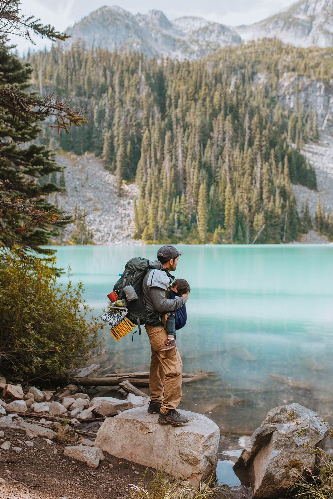 Person with a child on their back, standing by a turquoise lake with mountains in the background